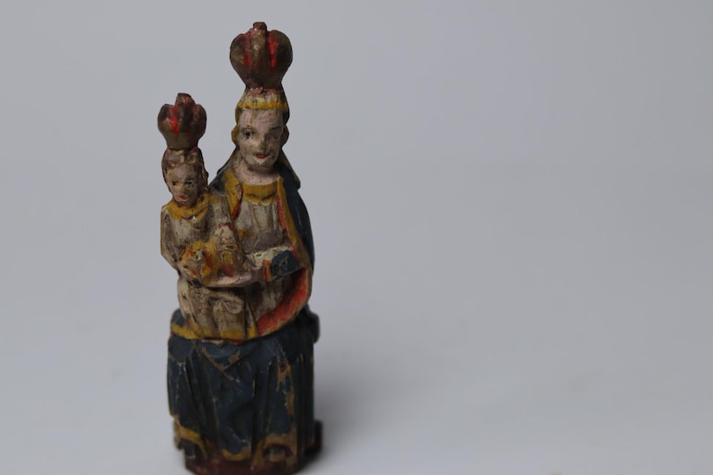 Pode incluir: Uma estatueta de madeira esculpida &agrave; m&atilde;o representando uma figura segurando uma crian&ccedil;a, ambas usando coroas. A escultura apresenta um acabamento de pintura desgastado em tons de azul, amarelo, vermelho e branco, com sinais de desgaste.