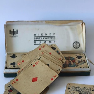 May include: A vintage deck of playing cards for Rummy, featuring aged, off-white card stock with red and black suits. The cards are partially fanned out, resting in an open cardboard box labeled WIENER SPIELKARTEN RUMMY with Austria branding.