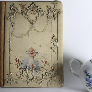 Op de afbeelding: Een antiek boek met een crèmekleurige kaft voorzien van reliëf-krulwerk, bloemenslingers en een centrale illustratie van een gevleugelde figuur die fluit speelt. Daarnaast staat een witte porseleinen theepot met blauwe bloemmotieven.