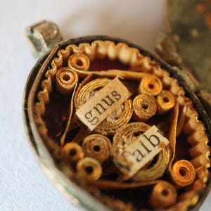 Pode incluir: Um close-up detalhado de um medalhão vintage aberto, revelando um diorama em miniatura. Elementos de papel enrolado e pequenas etiquetas com as palavras "gnus" e "albi" são visíveis. O medalhão tem uma moldura de metal dourado.