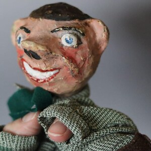 Könnte beinhalten: Eine Vintage-Puppe mit bemaltem Gesicht, blauen Augen und einem breiten, rotlippigen Lächeln. Die Puppe trägt eine grüne Fliege und eine Tweedjacke. Das Gesicht der Puppe weist Gebrauchsspuren auf, was ihren Charme ausmacht.