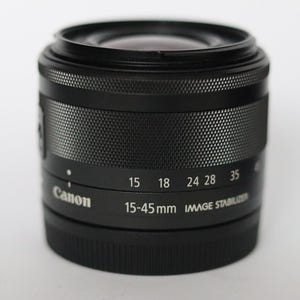 Pode incluir: Lente Canon preta 15-45mm com estabilizador de imagem. A lente tem uma pega texturizada e marcações de distância focal: 15, 18, 24, 28 e 35 mm. O logótipo Canon é visível.