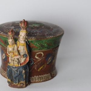 Pode incluir: Uma caixa redonda de madeira castanha com tampa, decorada com motivos florais pintados &agrave; m&atilde;o em azul, amarelo e verde. Na parte frontal, est&aacute; fixada uma figura de madeira esculpida de uma pessoa coroada com uma crian&ccedil;a, pintada em azul, amarelo e vermelho.