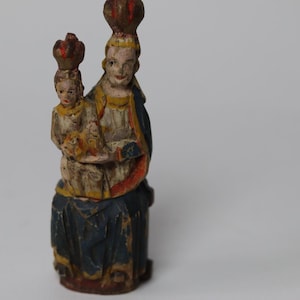 Pode incluir: Uma estatueta de madeira esculpida &agrave; m&atilde;o representando uma figura sentada segurando uma crian&ccedil;a, ambas usando coroas. A escultura apresenta detalhes pintados em azul, branco, amarelo e vermelho, mostrando sinais de desgaste e idade.