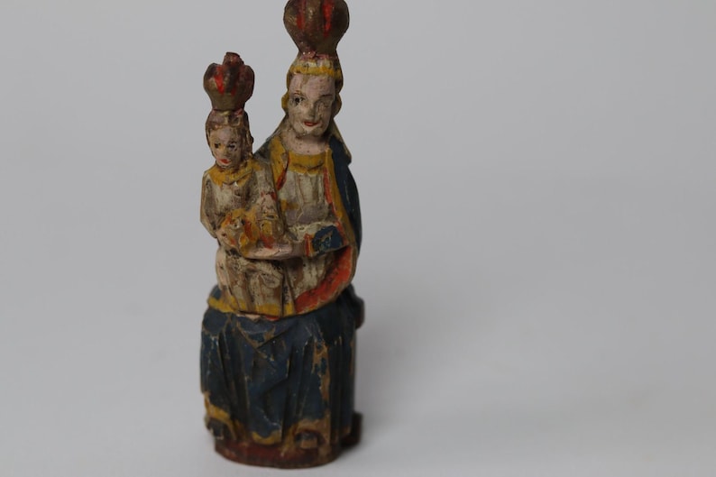 Pode incluir: Uma estatueta de madeira esculpida &agrave; m&atilde;o representando uma figura sentada segurando uma crian&ccedil;a, ambas usando coroas. A escultura apresenta detalhes pintados em azul, branco, amarelo e vermelho, mostrando sinais de desgaste e idade.