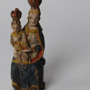 Pode incluir: Uma estatueta de madeira pintada &agrave; m&atilde;o representando uma figura segurando uma figura menor, ambas usando coroas. A figura maior veste uma t&uacute;nica azul-escura com detalhes em vermelho e amarelo, enquanto a figura menor &eacute; pintada em tons creme.