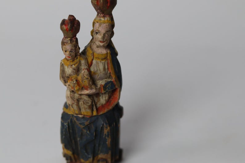 Pode incluir: Uma estatueta de madeira pintada &agrave; m&atilde;o representando uma figura segurando uma figura menor, ambas usando coroas. A figura maior veste uma t&uacute;nica azul-escura com detalhes em vermelho e amarelo, enquanto a figura menor &eacute; pintada em tons creme.