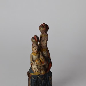 Pode incluir: Uma pequena estatueta de madeira pintada &agrave; m&atilde;o representando uma figura sentada segurando uma crian&ccedil;a. Ambas as figuras usam coroas e est&atilde;o vestidas com mantos pintados em tons de azul, vermelho e amarelo. A pintura mostra sinais de desgaste.