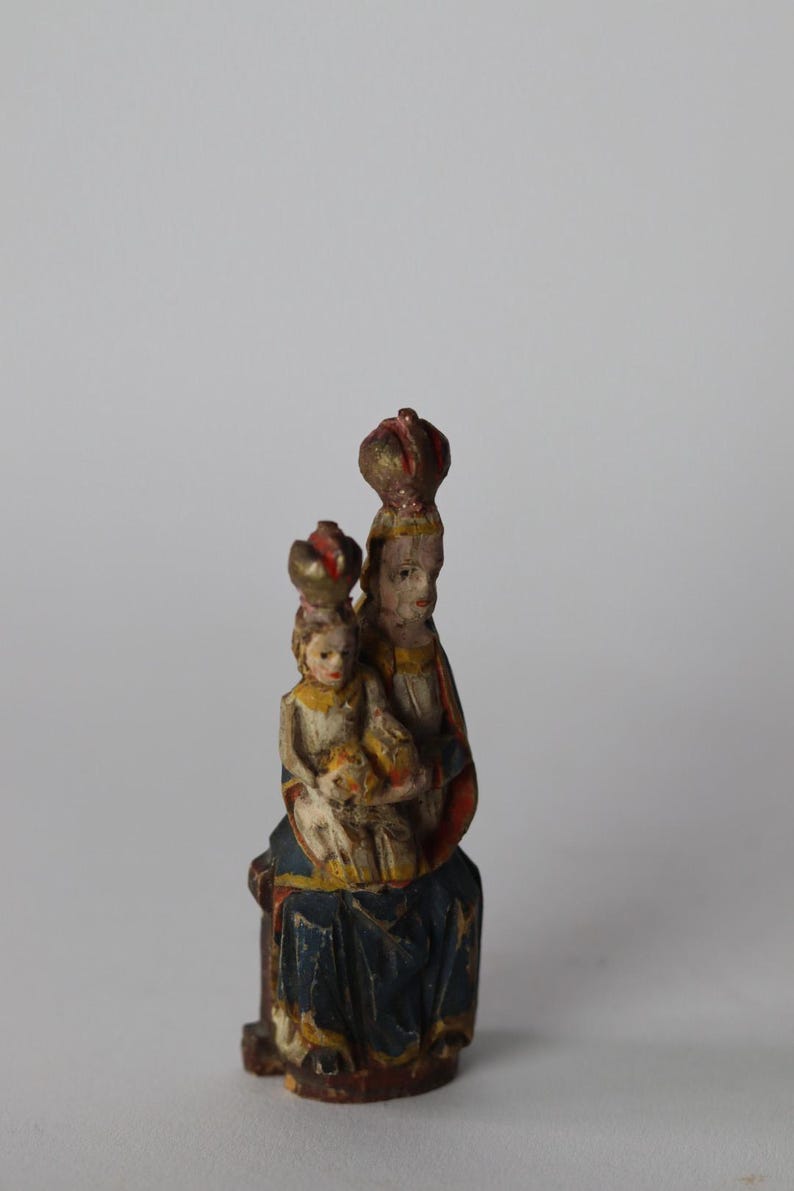 Pode incluir: Uma pequena estatueta de madeira pintada &agrave; m&atilde;o representando uma figura sentada segurando uma crian&ccedil;a. Ambas as figuras usam coroas e est&atilde;o vestidas com mantos pintados em tons de azul, vermelho e amarelo. A pintura mostra sinais de desgaste.