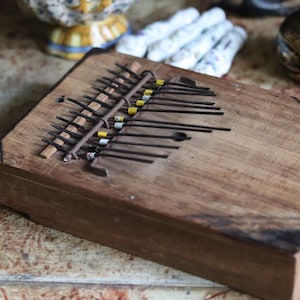 May include: A rectangular wooden kalimba featuring a series of thin metal tines arranged across the top surface. Several of the metal tines are marked with small yellow and white bands. The wooden body has a natural brown finish with dark edges.