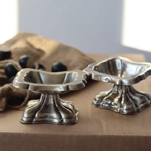 Può includere: Due ciotole a piedistallo argentate e ornate con bordo smerlato. Le ciotole sono su una superficie marrone con un panno di juta e diversi oggetti scuri e rotondi sullo sfondo. Le ciotole hanno una base decorativa e scanalata.