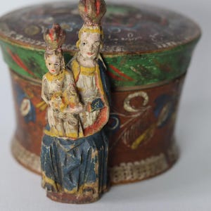Pode incluir: Uma caixa redonda de madeira com tampa, apresentando uma base castanha pintada &agrave; m&atilde;o com motivos florais em azul, vermelho e amarelo. Na parte frontal, est&aacute; fixada uma figura de madeira esculpida de um adulto coroado segurando uma crian&ccedil;a.