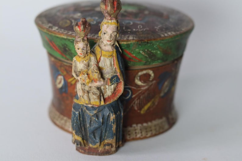 Pode incluir: Uma caixa redonda de madeira com tampa, apresentando uma base castanha pintada &agrave; m&atilde;o com motivos florais em azul, vermelho e amarelo. Na parte frontal, est&aacute; fixada uma figura de madeira esculpida de um adulto coroado segurando uma crian&ccedil;a.