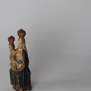 Pode incluir: Uma pequena estatueta de madeira pintada &agrave; m&atilde;o representando uma figura sentada segurando uma figura menor, ambas usando coroas. A escultura apresenta um manto azul-escuro com detalhes amarelos e sinais vis&iacute;veis de tinta envelhecida.