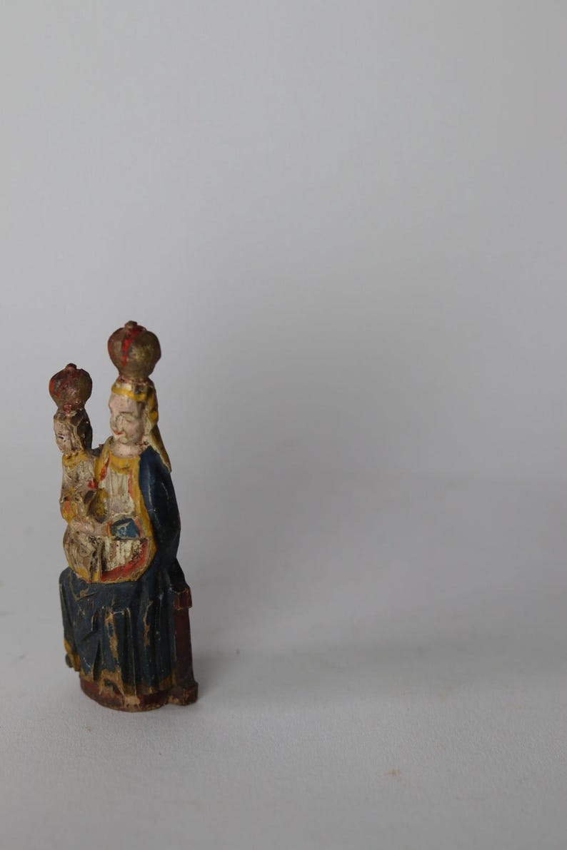 Pode incluir: Uma pequena estatueta de madeira pintada &agrave; m&atilde;o representando uma figura sentada segurando uma figura menor, ambas usando coroas. A escultura apresenta um manto azul-escuro com detalhes amarelos e sinais vis&iacute;veis de tinta envelhecida.