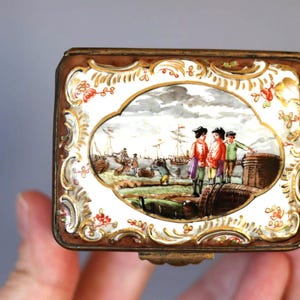 May include: An antique rectangular trinket box with a hand-painted harbour scene. The box has a gold-coloured trim with floral accents and a hinged lid. The scene depicts figures in colourful clothing, with ships in the background.