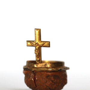 Op de afbeelding: Een goudkleurig kruisbeeld met een gedetailleerde afbeelding van Jezus Christus staat op een kleine, sierlijke container. De container staat op een bruine, getextureerde basis, waarschijnlijk van hout. De achtergrond is effen wit.