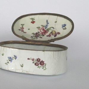 May include: An oval-shaped white enamel trinket box with a hinged lid and a metallic rim. The exterior and the interior of the lid feature hand-painted floral motifs in shades of pink, blue, red, and green, with a visible crackled texture.