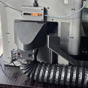 May include: A close-up of the back of a 3D printer. The printer is silver with black components, including a fan, a flexible exhaust hose, and a black funnel-shaped piece. A small orange component is visible.