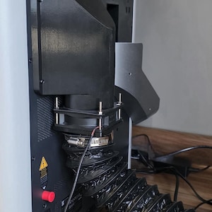 Könnte beinhalten: Ein schwarzer, 3D-gedruckter Abluftadapter an der Rückseite eines Geräts, mit einem quadratischen Lüftergehäuse, das mit Metallschrauben befestigt ist, und einem flexiblen, gewellten schwarzen Schlauch mit einer silbernen Schlauchschelle.