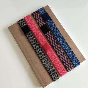 May include: Four elastic bands with distinct patterns arranged diagonally on a rectangular brown cardboard backing. The patterns include a black and white geometric print, a red hexagonal design, a blue and brown arrow motif, and a blue wave pattern.