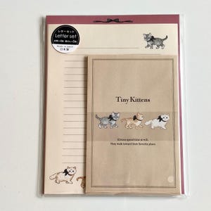 Puede incluir: Un juego de papelería en una funda de plástico transparente que incluye un sobre beige con el texto Tiny Kittens y tres gatitos ilustrados con lazos negros. El papel rayado incluye una pequeña ilustración de un gatito y un lazo.