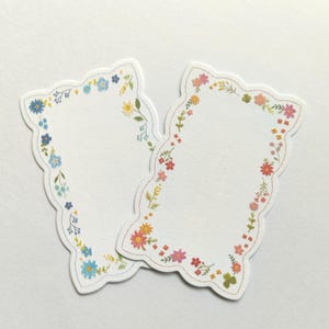 May include: Two die-cut paper labels with scalloped edges. The left label features a border of blue, yellow, and green floral illustrations. The right label features a border of pink, orange, yellow, and green floral illustrations on white paper.