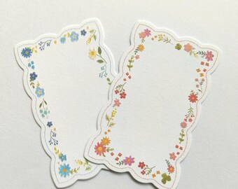 Puede incluir: Dos etiquetas de papel troqueladas con bordes festoneados. La etiqueta izquierda presenta un borde de ilustraciones florales azules, amarillas y verdes. La etiqueta derecha presenta un borde de flores rosas, naranjas, amarillas y verdes.