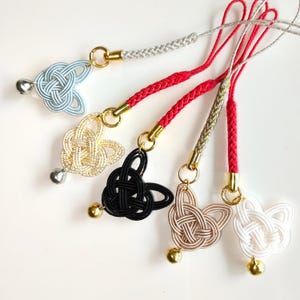 May include: Five decorative charms featuring intricate knotted cord designs in light blue, gold, black, beige, and white. Each charm is attached to a braided cord strap with a small metallic bell at the bottom and a gold-toned metal ring connector.