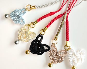Puede incluir: Cinco colgantes decorativos con intrincados diseños de cordón anudado en azul claro, dorado, negro, beige y blanco. Cada colgante está unido a una correa de cordón trenzado con una pequeña campana metálica y un anillo conector dorado.