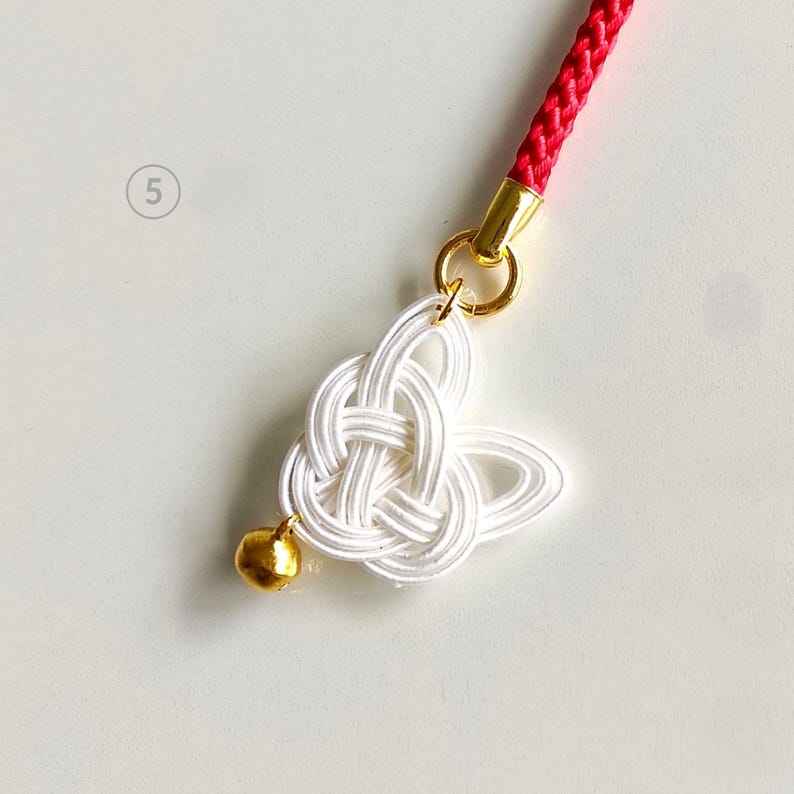 May include: A white mizuhiki knot charm shaped like a butterfly, attached to a red braided cord by a gold-colored metal ring. A small, round, gold-colored bell is attached to the bottom of the white cord knot. The number 5 is visible.