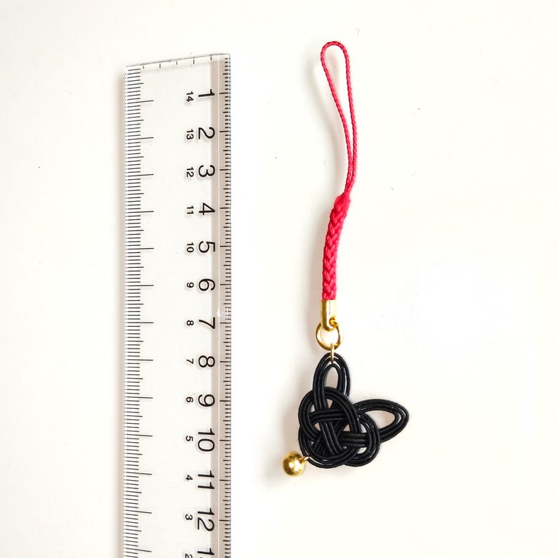 May include: A black cord charm featuring a knotted design and a small gold-colored bell, attached to a red braided loop. A clear plastic ruler is positioned next to the charm, showing the total length is approximately 5.5 inches from top to bottom.