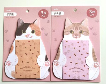 May include: Two packs of three cat-shaped paper envelopes on pink backing cards. Each envelope features a cat illustration holding a rectangular pouch printed with paw prints, fish icons, and the text Nyan Nyan Crew, Cats love freedom and friends.