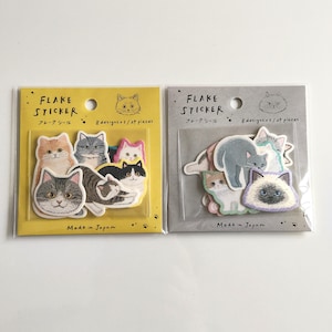 May include: Two packages of flake stickers featuring cat illustrations. The yellow package and gray package each contain 24 pieces with 8 designs. The text on the packaging reads Flake Sticker, 8 designs x 3 / 24 pieces, and Made in Japan.
