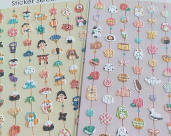 Puede incluir: Dos hojas de pegatinas Sticker Selection con filas de pequeños iconos texturizados de temática japonesa, incluyendo gatos, comida, flores y símbolos tradicionales, dispuestos en líneas verticales sobre fondos amarillo claro y morado claro.