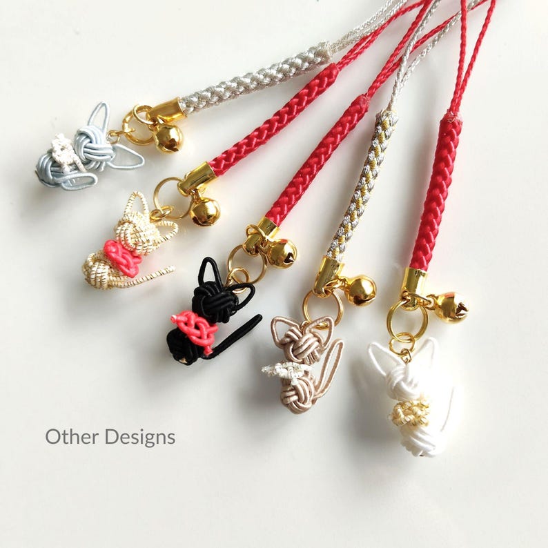 May include: Five decorative charms featuring cat-shaped figures crafted from knotted cord in light blue, beige, black, tan, and white. Each charm includes a small gold-toned bell and a braided cord strap in red or silver. The text Other Designs is visible.