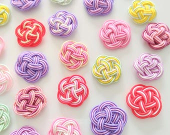 May include: A collection of decorative flower-shaped knots made from colorful cord. The knots feature intricate woven patterns in various shades of pink, purple, yellow, red, and light green, with some designs incorporating white accent strands.