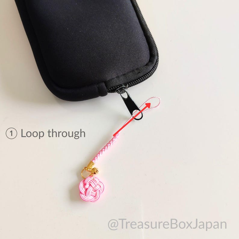 May include: A black zippered case with a pink braided cord strap attached to the zipper pull. The strap features a small pink knotted charm and a gold-colored metal ring. Text on the image reads: 1 Loop through and @TreasureBoxJapan.