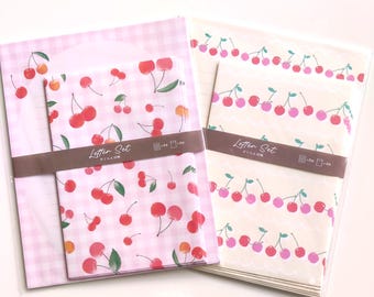 Puede incluir: Dos juegos de papelería en envases de plástico transparente. El conjunto de la izquierda presenta un patrón de cuadros vichy rosa con ilustraciones de cerezas rojas. El de la derecha tiene un fondo crema con filas de cerezas.
