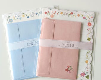 Puede incluir: Dos juegos de papelería en envases de plástico transparente. Un juego incluye un sobre y papel azul claro con un borde floral azul. El otro juego tiene un sobre rosa con un pequeño detalle floral y papel con un borde de flores de colores.