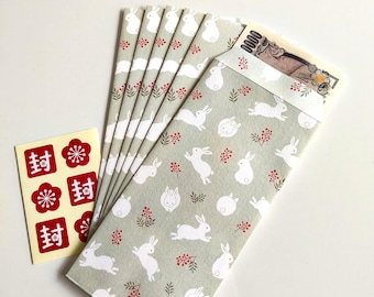 Puede incluir: Un juego de sobres de papel verde claro con un patrón repetitivo de conejos blancos y pequeñas bayas rojas, uno de los cuales contiene un billete japonés de 10.000 yenes. Se incluye una pequeña hoja de pegatinas con sellos rojos y flores.