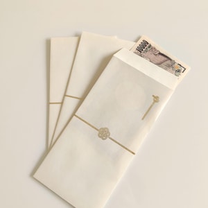 Könnte beinhalten: Drei cremefarbene, rechteckige Papierumschläge mit einer horizontalen goldenen Linie und einem zentralen goldenen Knotendesign. Ein Umschlag ist oben leicht geöffnet und zeigt einen japanischen 10000-Yen-Schein mit dem Text 10000.