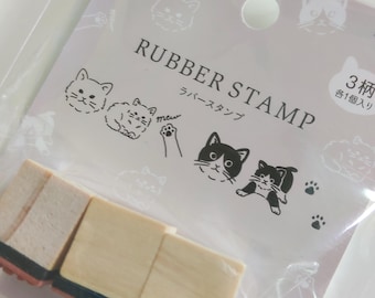 Puede incluir: Un juego de tres sellos de goma con bloques de madera clara, empaquetados en una funda de plástico transparente. El empaque presenta el texto RUBBER STAMP, caracteres japoneses e ilustraciones de gatos y huellas de gato.