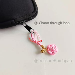 May include: A black zipper pull attached to a black case, featuring a pink braided cord loop and a pink knotted charm with a small gold bell. The image includes the text 2 Charm through loop and @TreasureBoxJapan with a red arrow graphic.