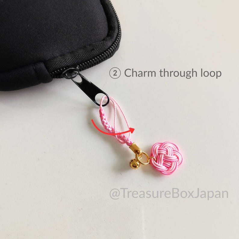 May include: A black zipper pull attached to a black case, featuring a pink braided cord loop and a pink knotted charm with a small gold bell. The image includes the text 2 Charm through loop and @TreasureBoxJapan with a red arrow graphic.