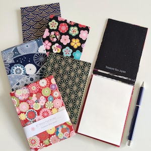 May include: A collection of B7 memo pad holders featuring various fabric covers with floral, geometric, and wave patterns. One holder is open to reveal white paper and the text Treasure Box Japan. A blue pen lies next to the memo pads.
