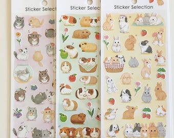 Puede incluir: Tres hojas verticales de pegatinas etiquetadas como Sticker Selection y Made in Japan. La hoja izquierda presenta chinchillas grises y marrones con flores y frutas. La hoja central muestra cobayas marrones y blancas con manzanas y tulipanes. La hoja derecha muestra varios conejos con fresas y flores.