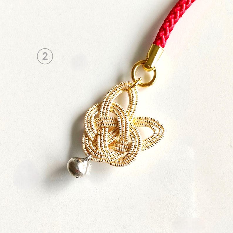May include: A decorative charm featuring a gold-colored knotted cord design attached to a bright red braided cord. The knot is accented with a small, smooth silver-colored bell at the bottom. A small circle with the number 2 is visible nearby.