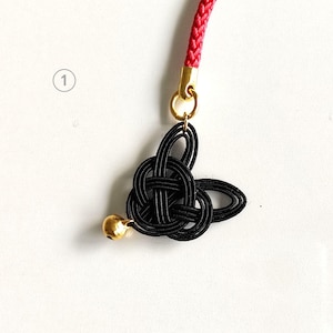 May include: A black mizuhiki knot charm featuring a small gold-toned bell, attached to a red braided cord by a gold-toned metal jump ring. The knot is shaped into a stylized form with multiple loops, and the number one is visible in a circle.