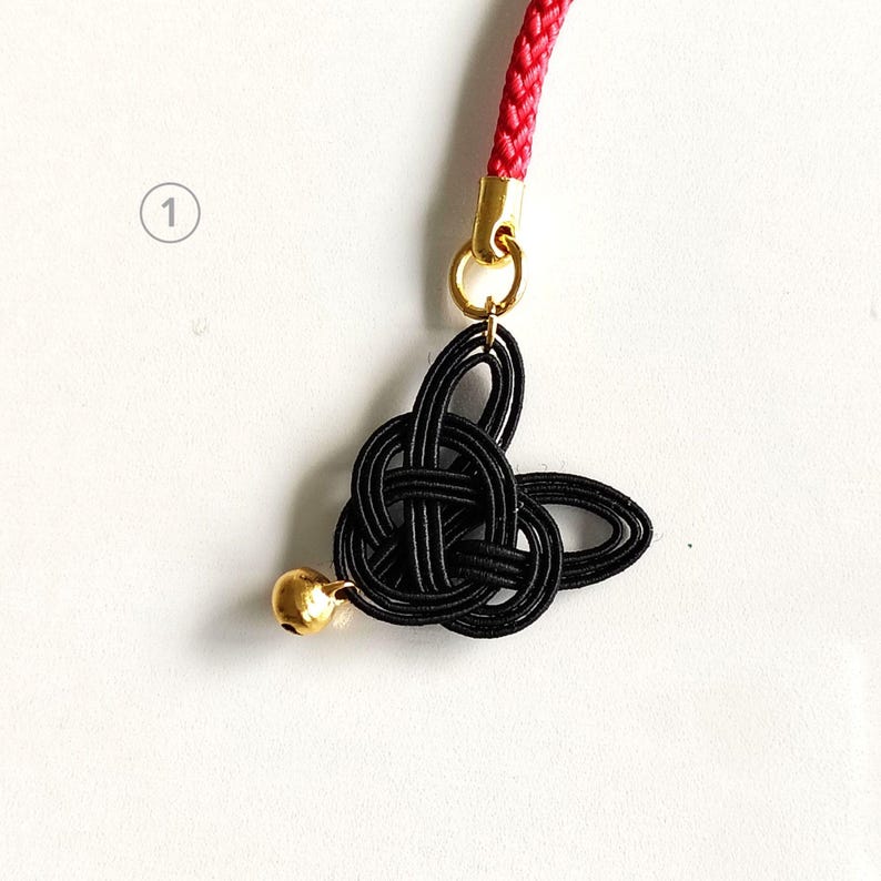 May include: A black mizuhiki knot charm featuring a small gold-toned bell, attached to a red braided cord by a gold-toned metal jump ring. The knot is shaped into a stylized form with multiple loops, and the number one is visible in a circle.